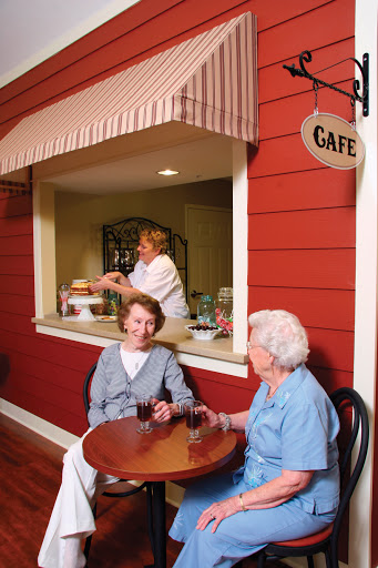 Two seniors enjoy a conversation over drinks at a cozy cafe, with a staff member serving from the counter.