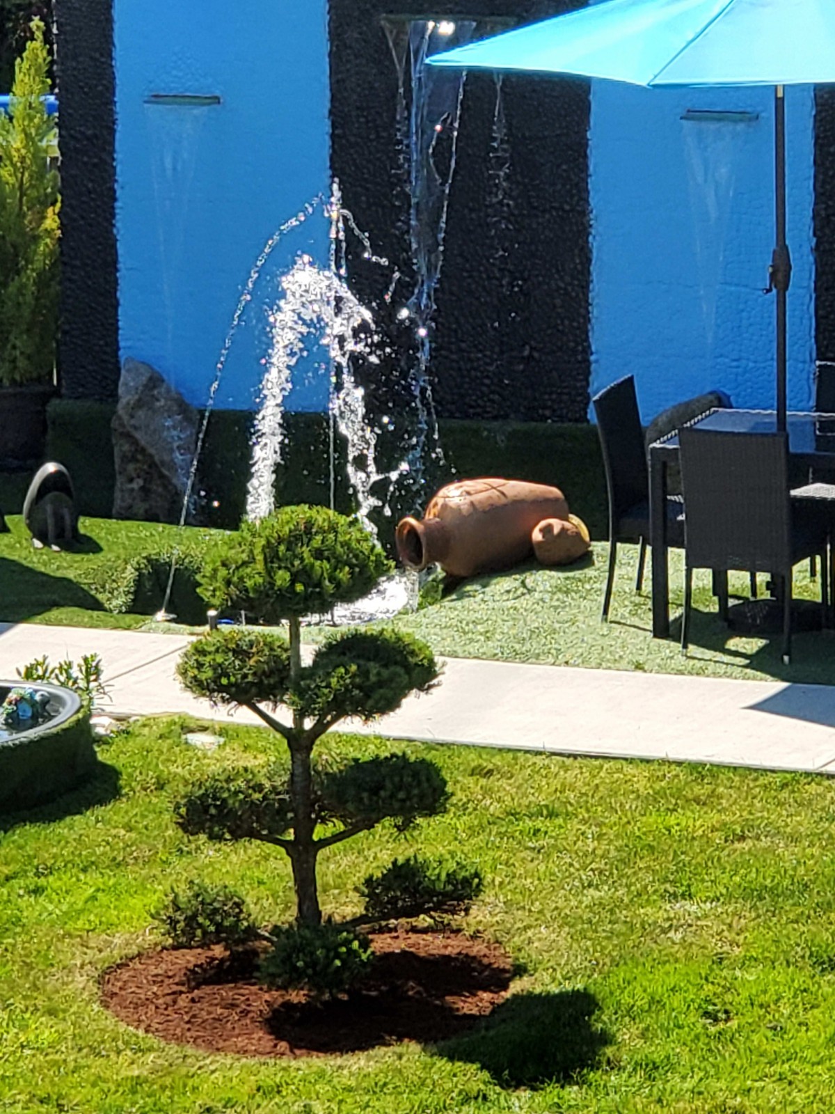 A serene garden setting with a fountain, neatly trimmed shrubs, and patio furniture on a sunny day.