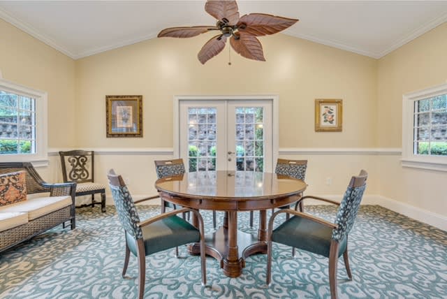 A cozy dining room featuring a round table with six chairs, a ceiling fan with leaf-shaped blades, and artwork adorning the walls.
