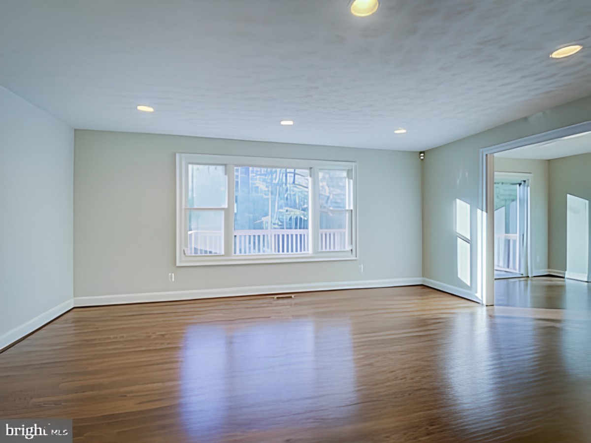 A spacious, empty room with hardwood flooring and a large window allowing natural light to fill the space.