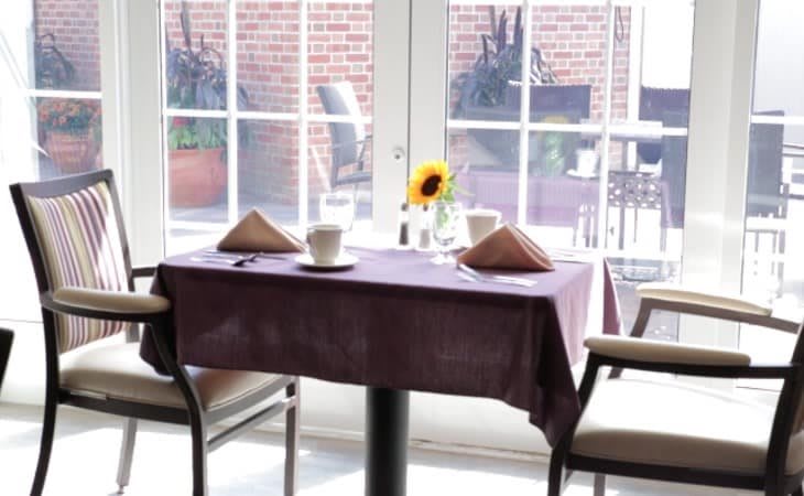 A neatly set dining table with a sunflower centerpiece by a large window.