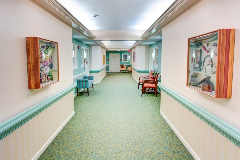 A well-lit hallway with carpeted flooring, featuring chairs and paintings on the walls.