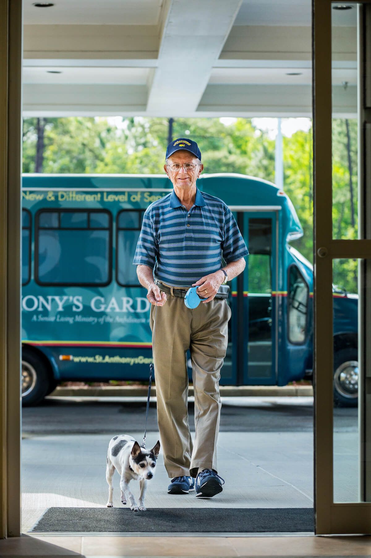 An elderly man in a striped shirt and hat walks with his dog near a bus.