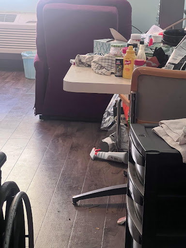 A cluttered sitting area featuring a table, wheelchair, and various personal items.