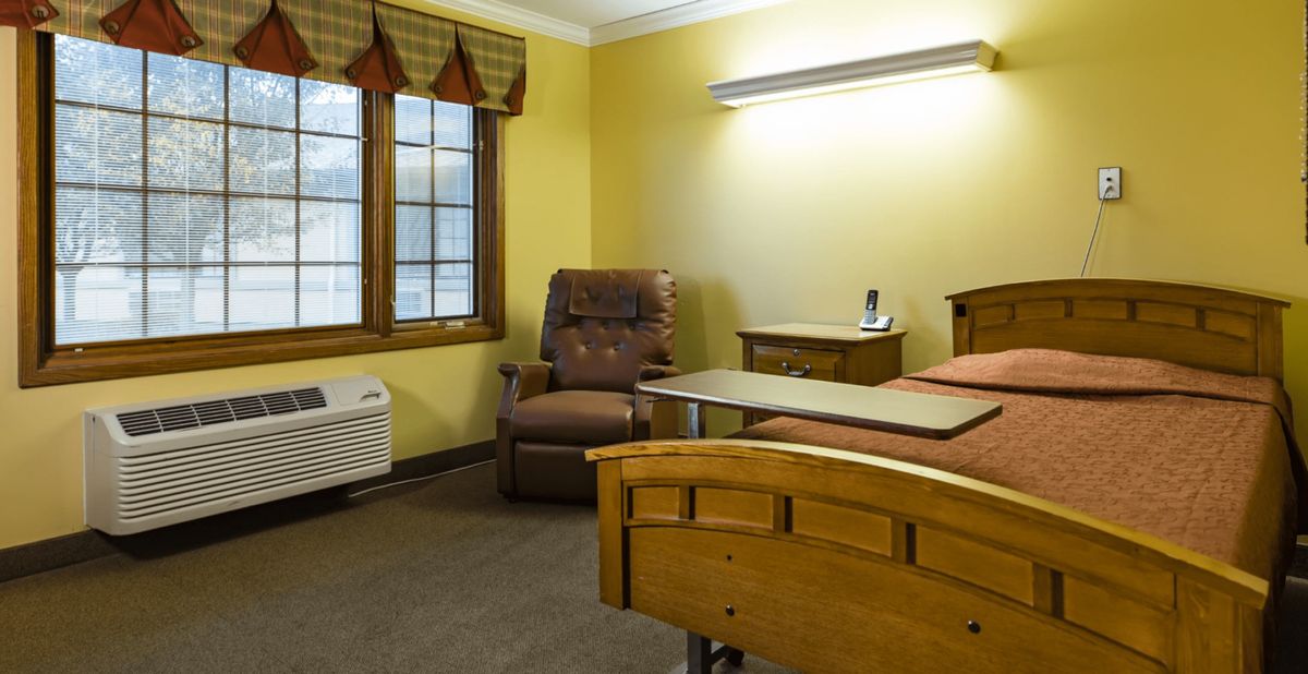 A cozy hospital room with a bed and recliner chair next to a window with yellow walls.