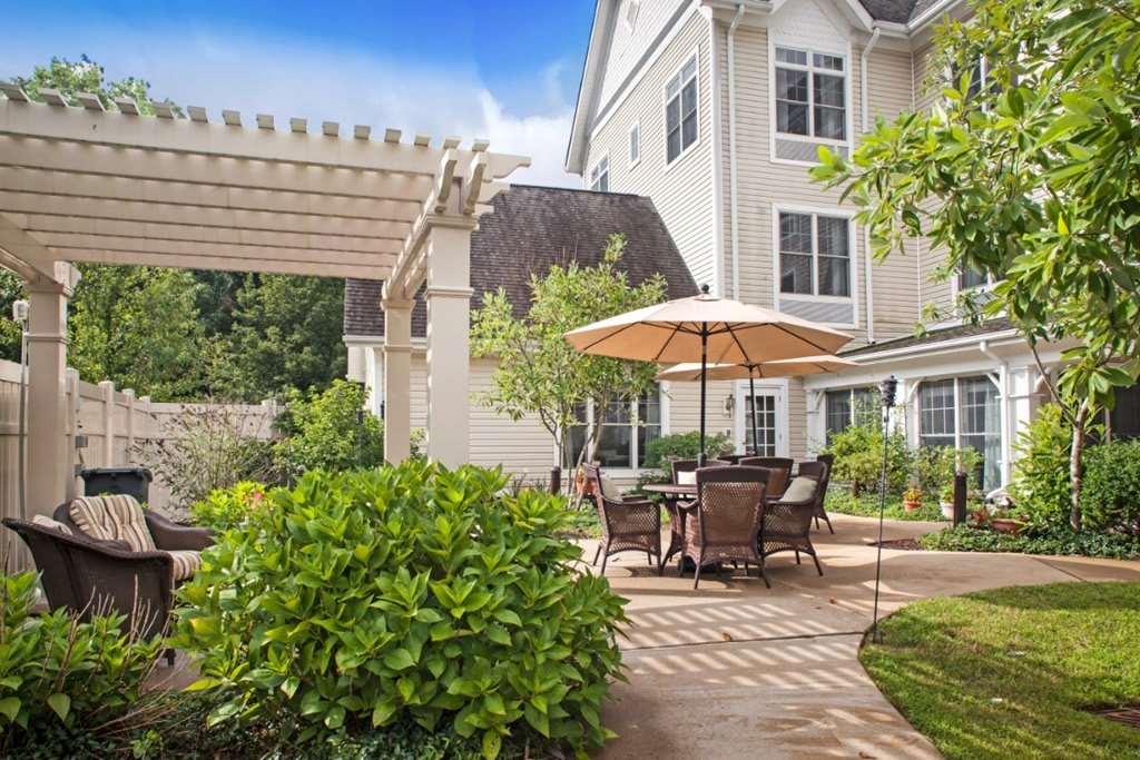 A sunny backyard patio with outdoor furniture and a garden umbrella, surrounded by lush greenery and connected to a large house.