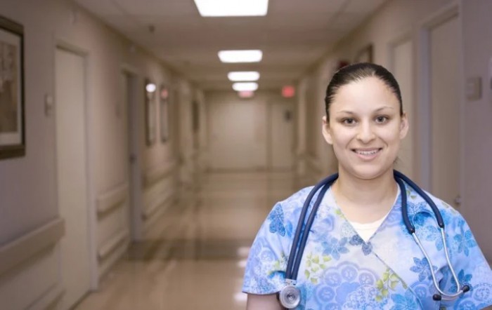 A nurse stands in a hospital hallway with a stethoscope around her neck.