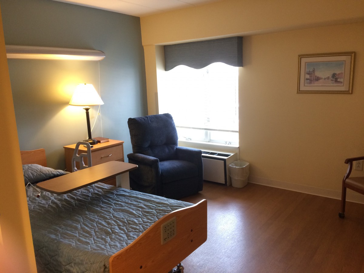 A simple hospital room with a bed, chair, and lamp near a window.