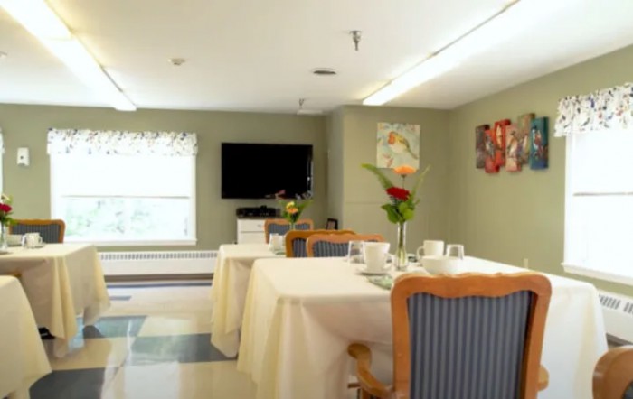 A dining room with neatly arranged tables and chairs, featuring a television and floral decorations.