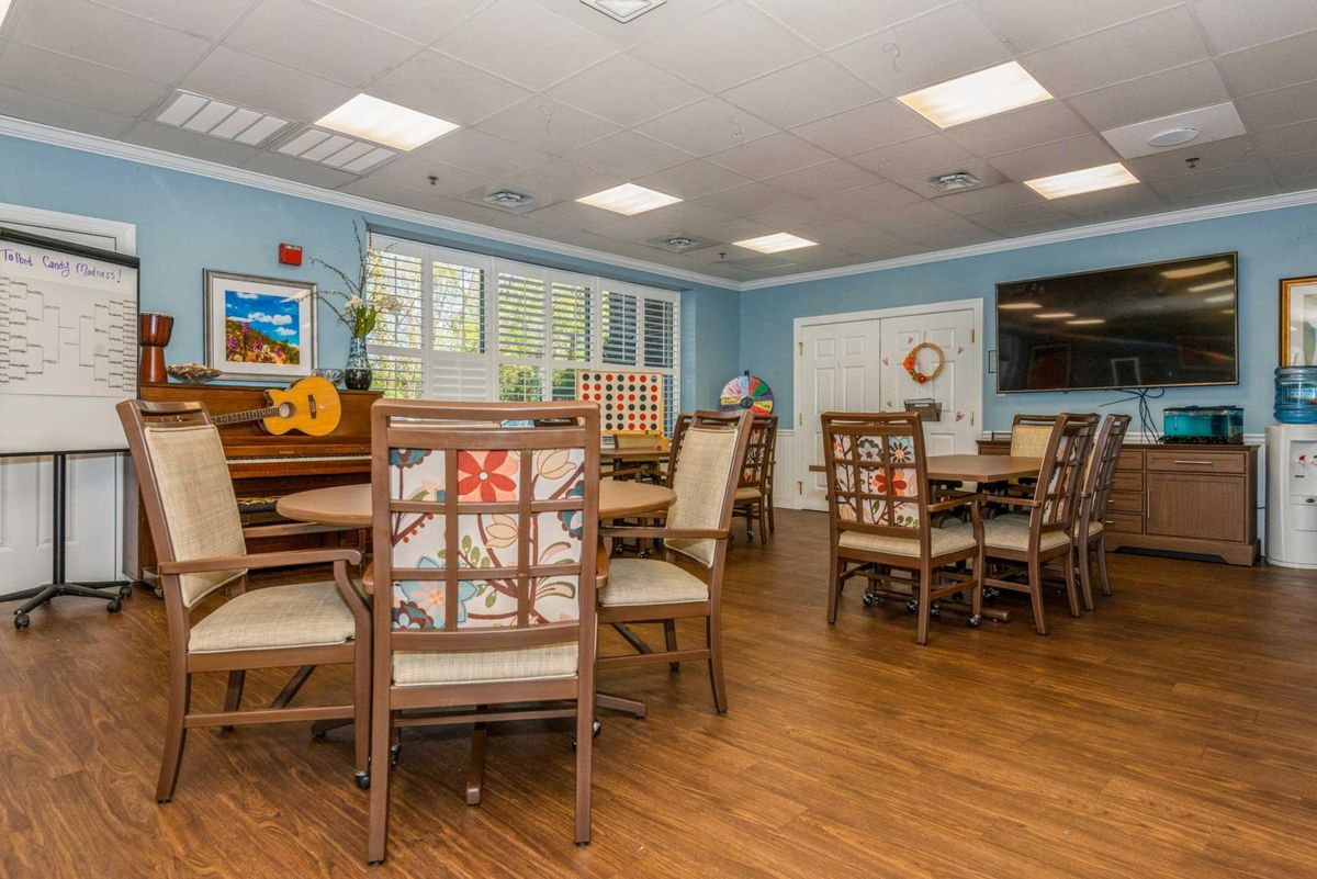 A spacious community room with tables, chairs, and a piano, featuring bright lighting and wooden flooring.