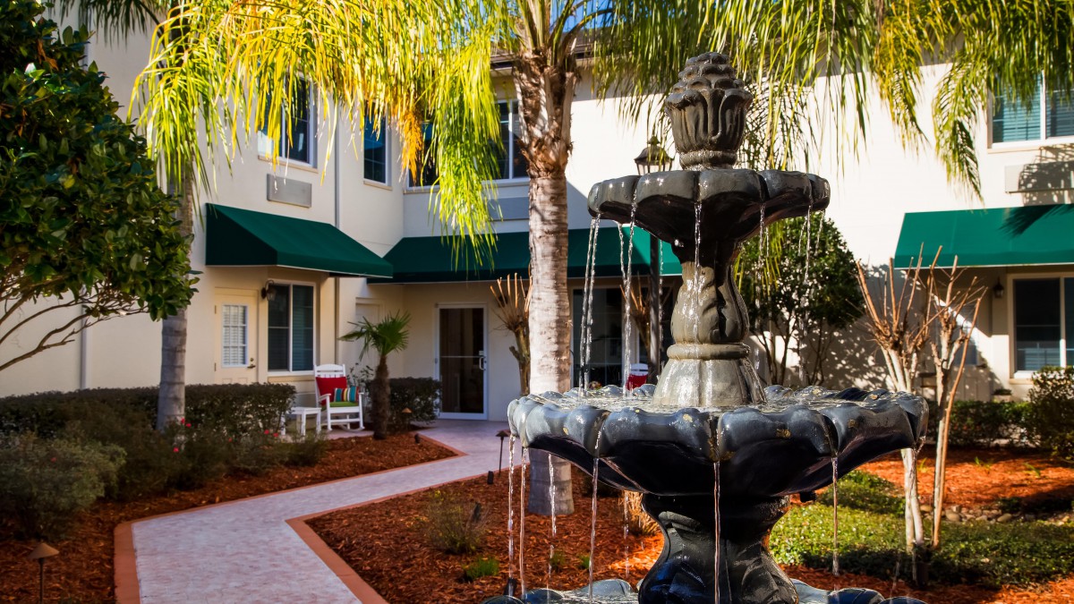 A tranquil courtyard featuring a decorative fountain surrounded by palm trees and a building with green awnings.