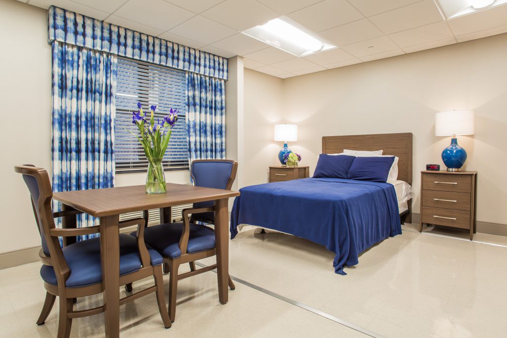 A neatly decorated bedroom featuring a blue color scheme, with a bed, table and chairs, and flowers in a vase by the window.