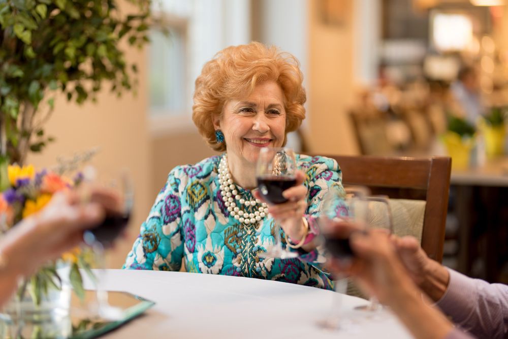 An elderly woman enjoying a social gathering with friends at a dining table, raising a glass of wine in celebration.