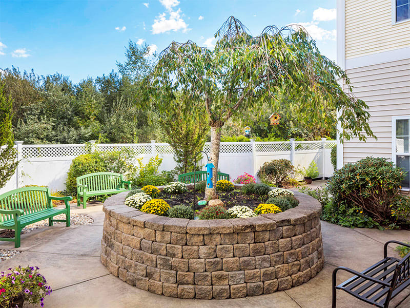 A beautifully landscaped patio with a circular stone planter featuring lush greenery and trees, set against a bright, clear day.
