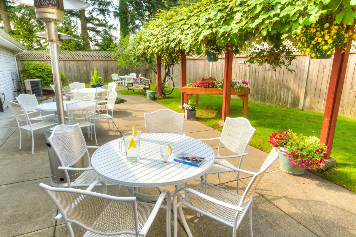 A sunny garden patio with white tables and chairs, surrounded by greenery.