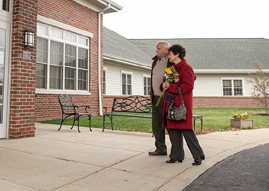 An elderly couple walking outside, carrying flowers near a building.