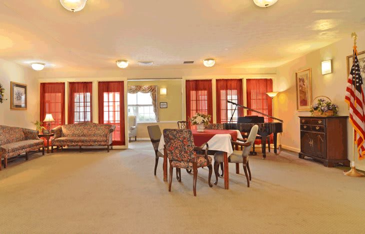 A spacious living room with a dining area and piano, featuring carpeted flooring and large windows with red curtains.