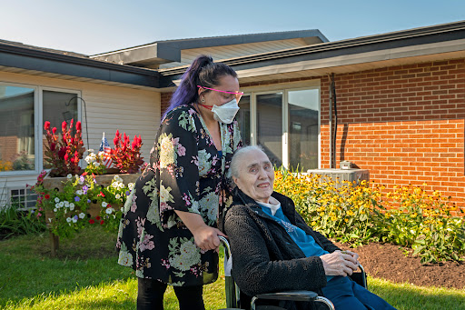 A caregiver assists an elderly person in a wheelchair, surrounded by blooming flowers on a sunny day.