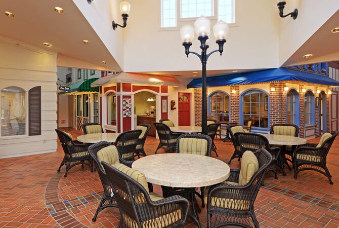 A cozy indoor atrium with tables, wicker chairs, and storefront facades under decorative street lamps.