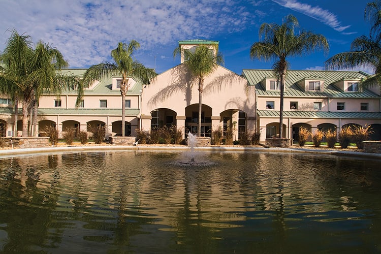 A scenic view of a building surrounded by palm trees, with a fountain reflecting in the water under a partly cloudy sky.