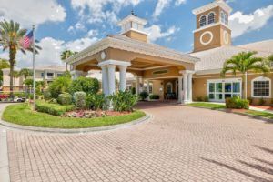 A picturesque entrance to a large building with a landscaped garden and partly cloudy sky.