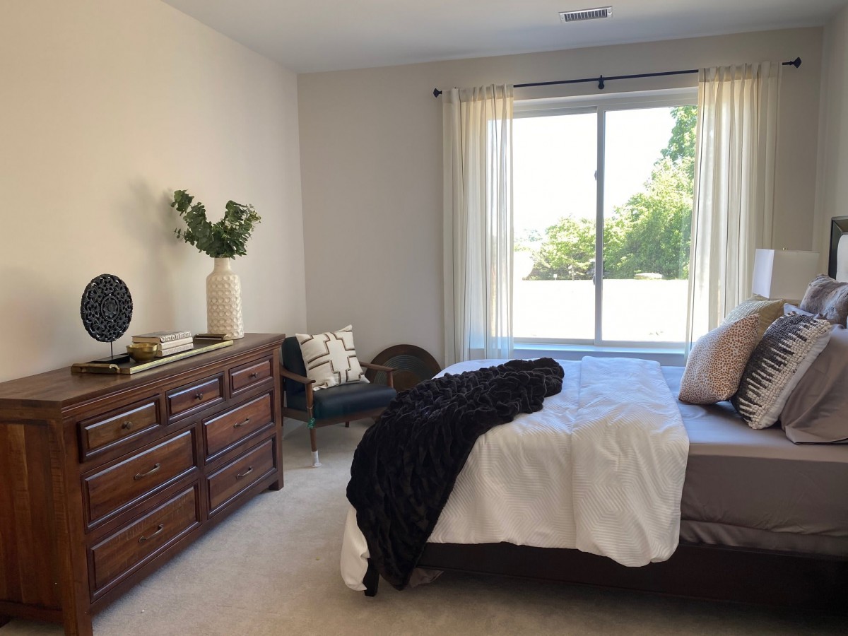 A minimalist bedroom with a large window allowing natural light, featuring a neatly made bed, a wooden dresser, and simple decor.