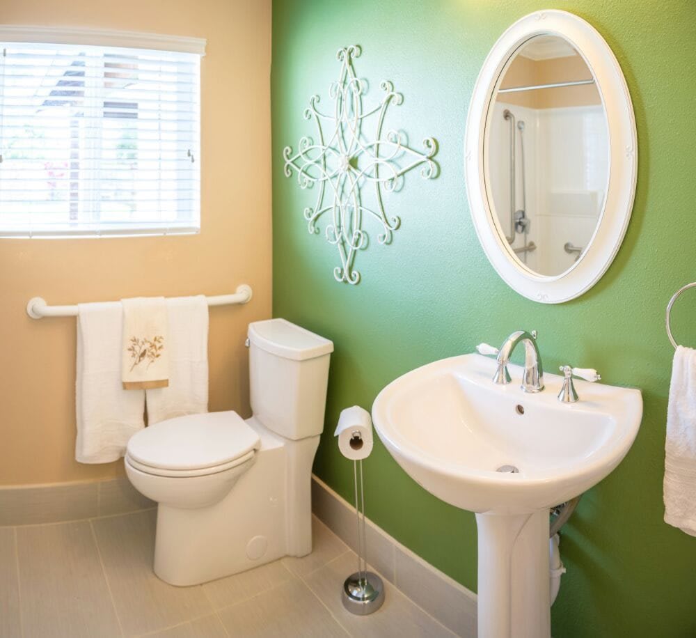 A bright and tidy bathroom featuring a pedestal sink, toilet, decorative wall art, and an oval mirror.