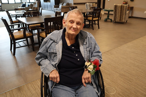 A smiling elderly woman in a wheelchair, adorned with a floral decoration, enjoying her time indoors.