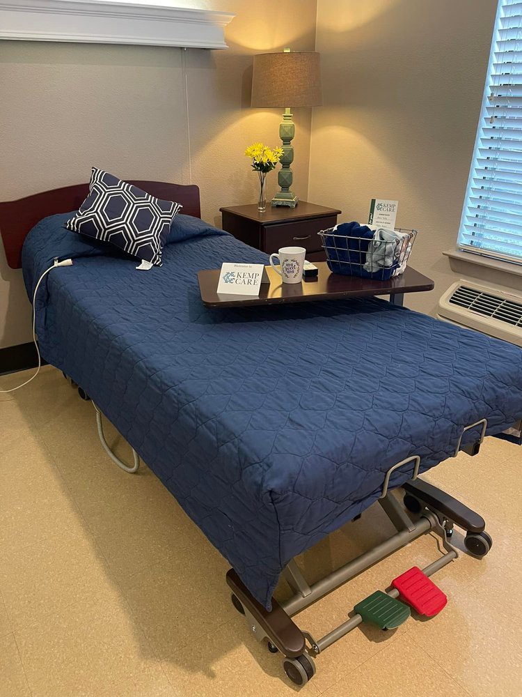 A hospital room with a neatly made bed featuring blue bedding and a decorative pillow, accompanied by a tray table topped with flowers and other items under a warm lamp.