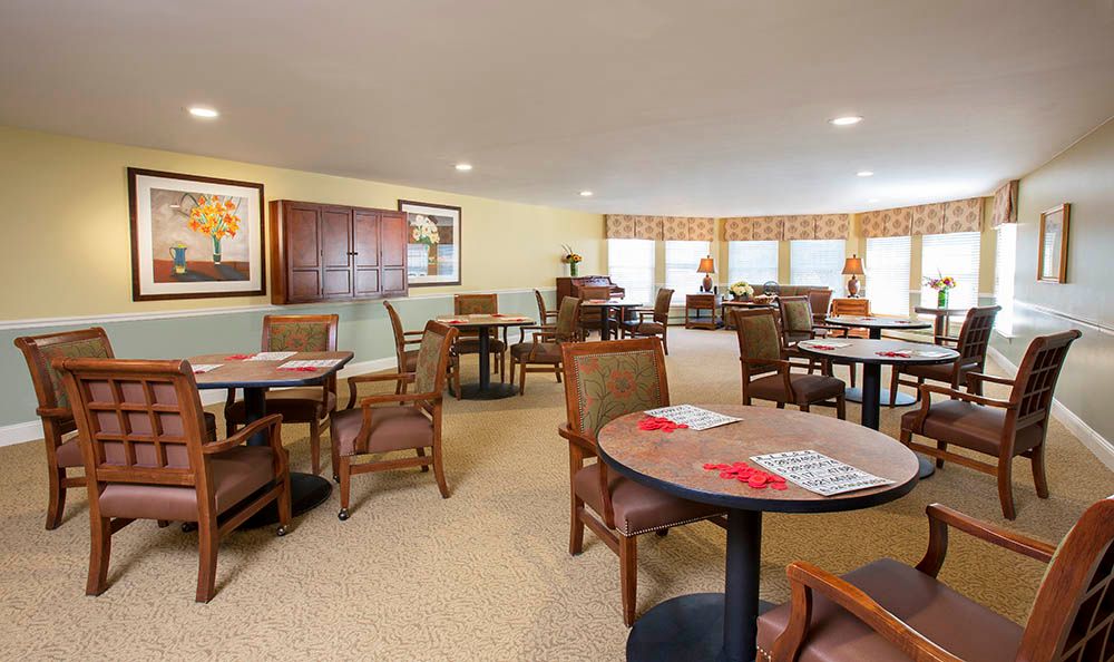 A cozy indoor seating area with multiple tables and chairs, featuring artwork on the walls and ample natural light from large windows.