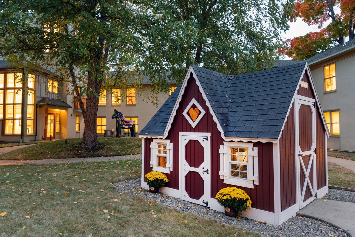 A quaint red playhouse sits in the yard, surrounded by trees and autumn decor, with a larger home visible in the background during an evening setting.