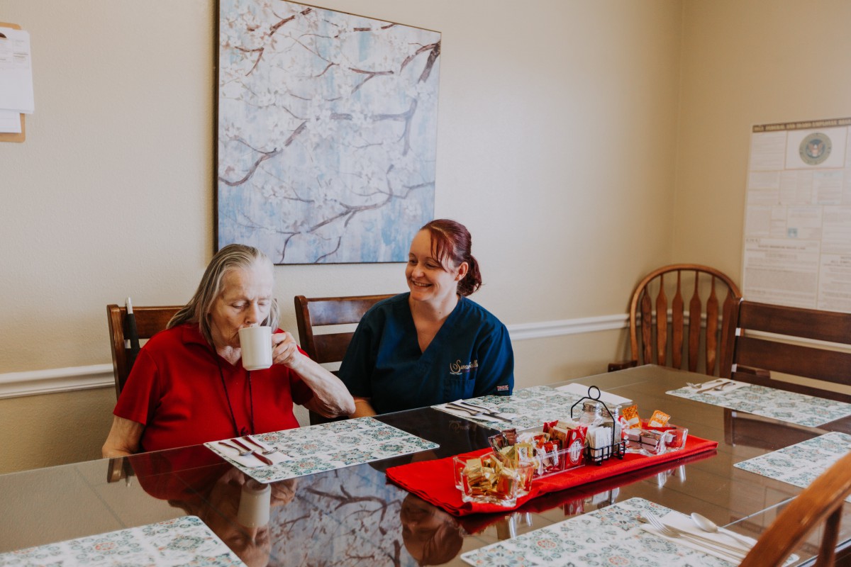 A caregiver assists a senior at a dining table.