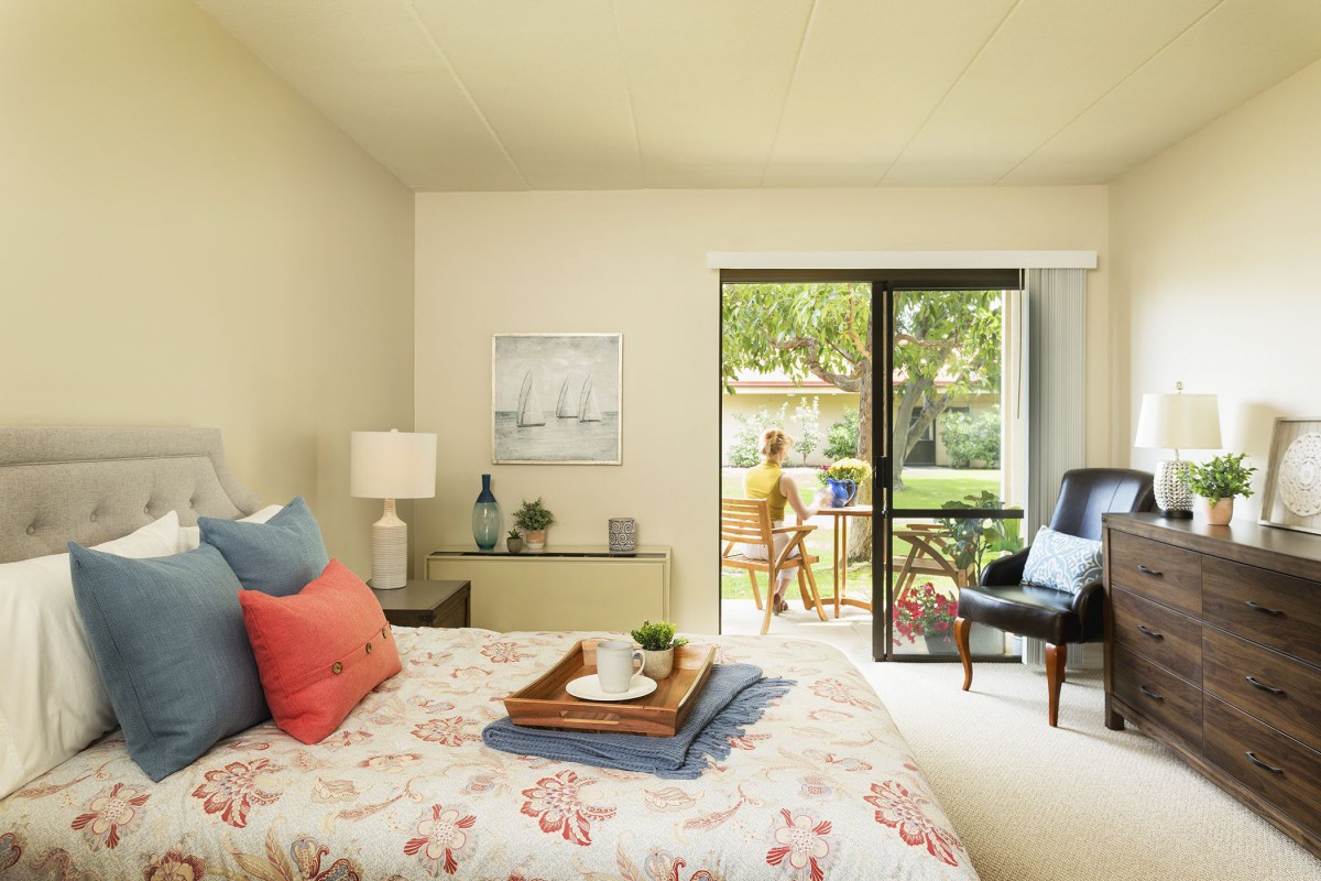 A bright and cozy bedroom with a bed adorned with pillows, a bedside lamp, and access to a balcony through sliding glass doors.