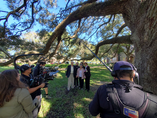 A media crew filming outdoors under a large tree, with several people posing for the camera.