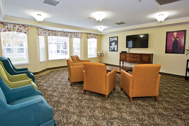A cozy living room with upholstered chairs facing a television, featuring large windows and patterned carpet.