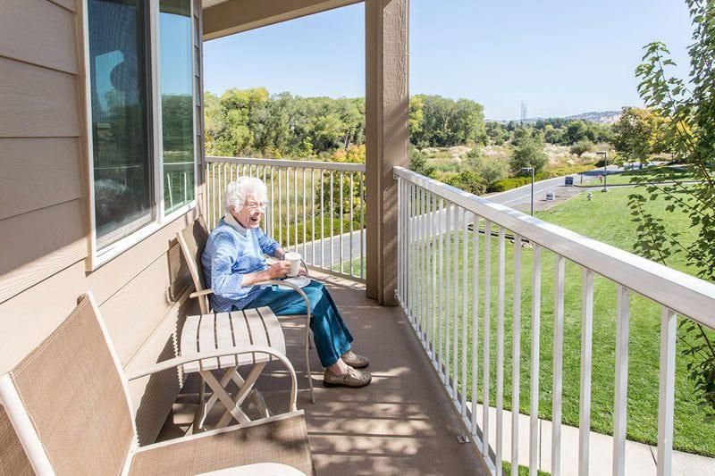 An elderly person sits on a sunny balcony, enjoying a book with a scenic view.