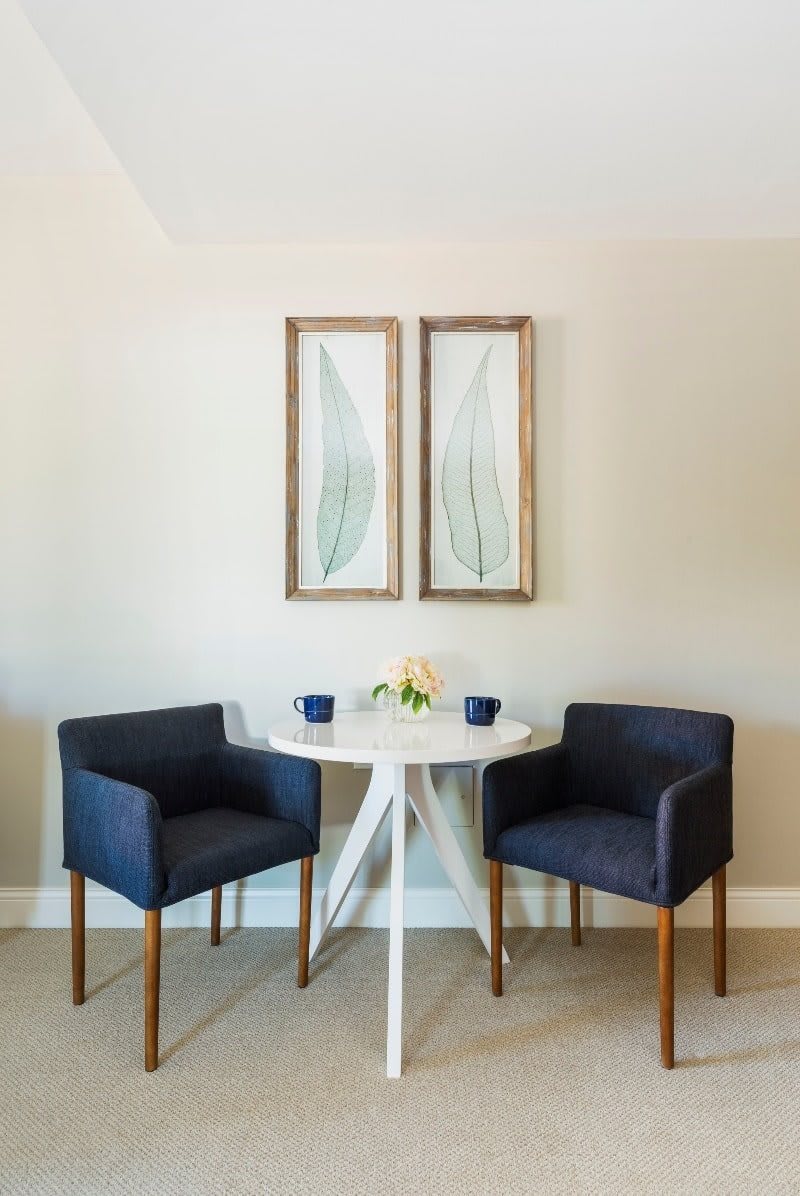 A modern minimalist dining area featuring a round white table, two navy chairs, and wall art.