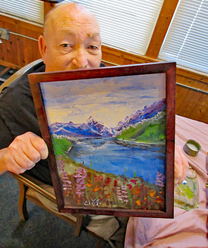 An elderly artist proudly displays their vibrant landscape painting featuring mountains and a serene lake.