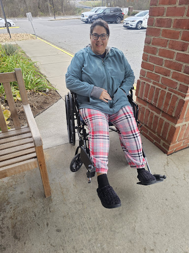 A woman sitting in a wheelchair outdoors, dressed in comfortable plaid pants and a casual jacket.