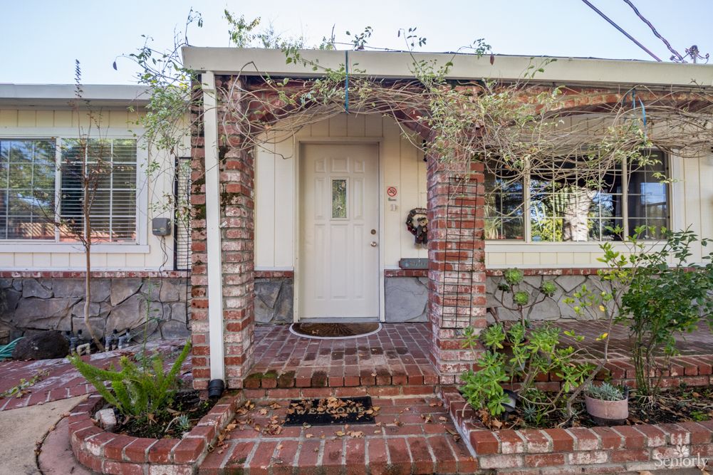 A house entrance with a brick archway covered in plants leading to a white front door.