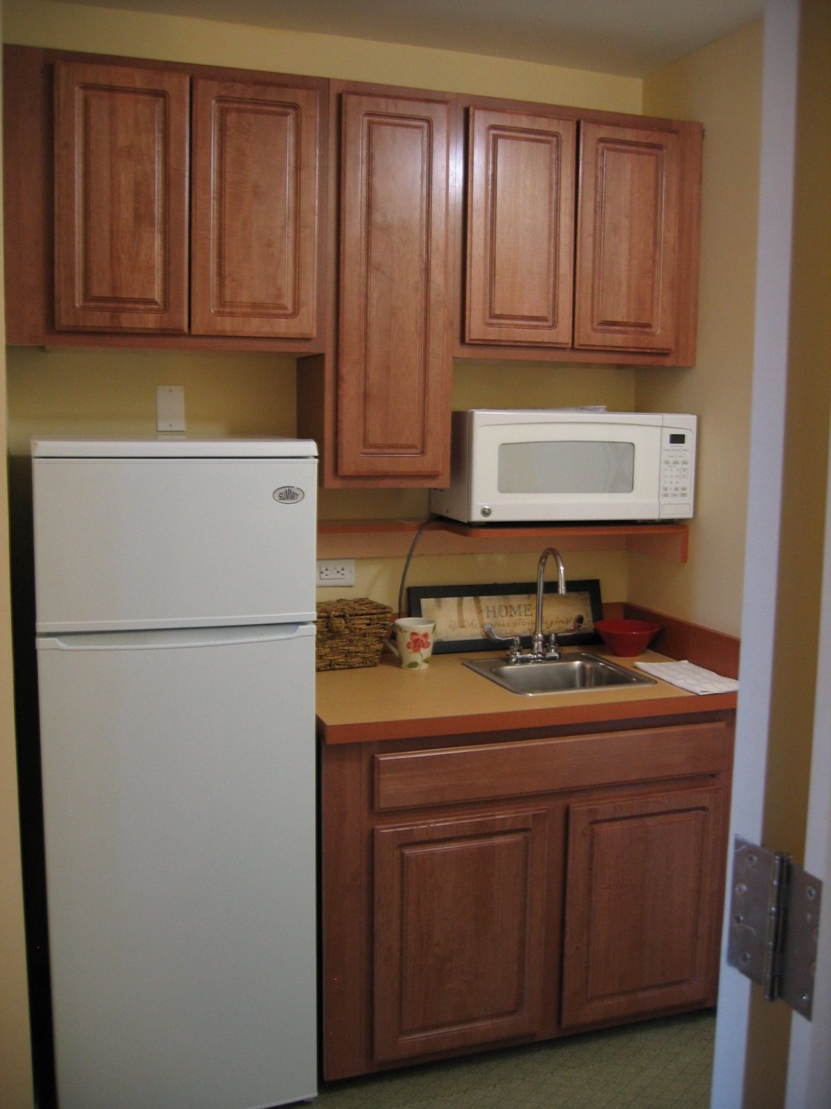 A compact kitchenette with wooden cabinets, a fridge, microwave, and sink.