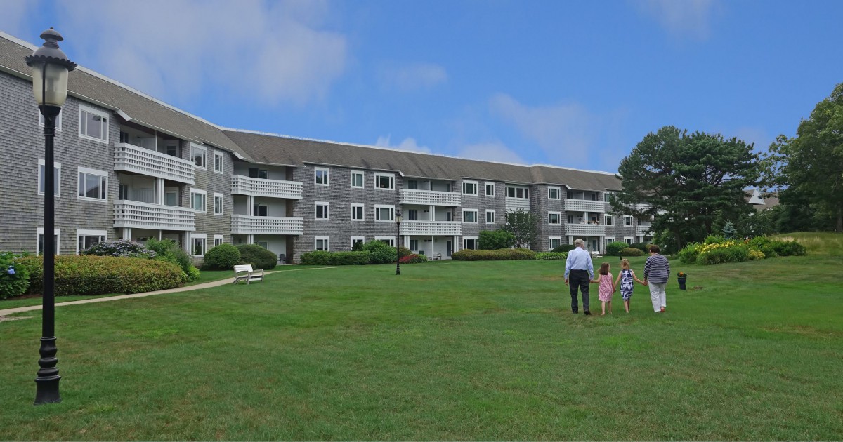 A group of people walking towards a large residential building on a grassy lawn under a blue sky.
