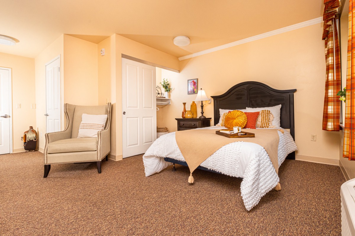 A cozy bedroom with warm colors, featuring a bed with orange and yellow pillows and a comfortable armchair.