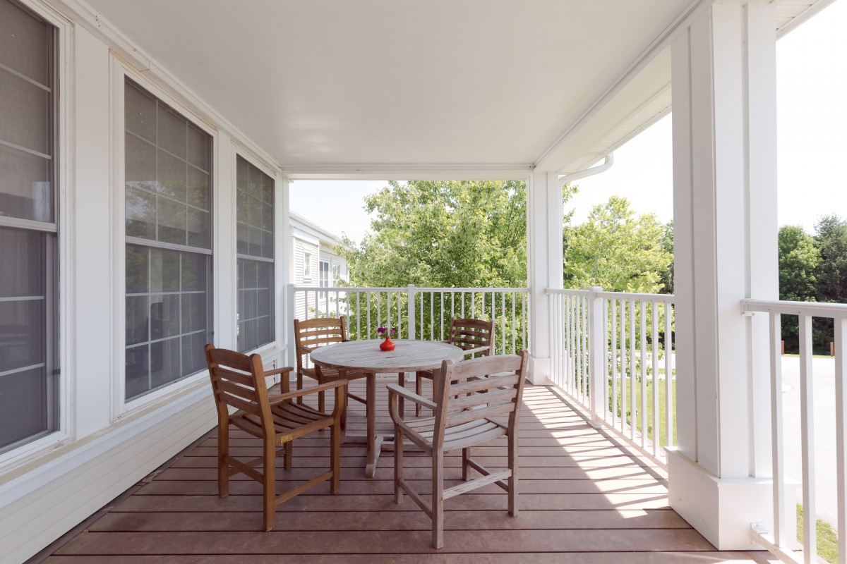 A covered porch with wooden deck flooring features a table and four chairs, surrounded by white railings on a sunny day.