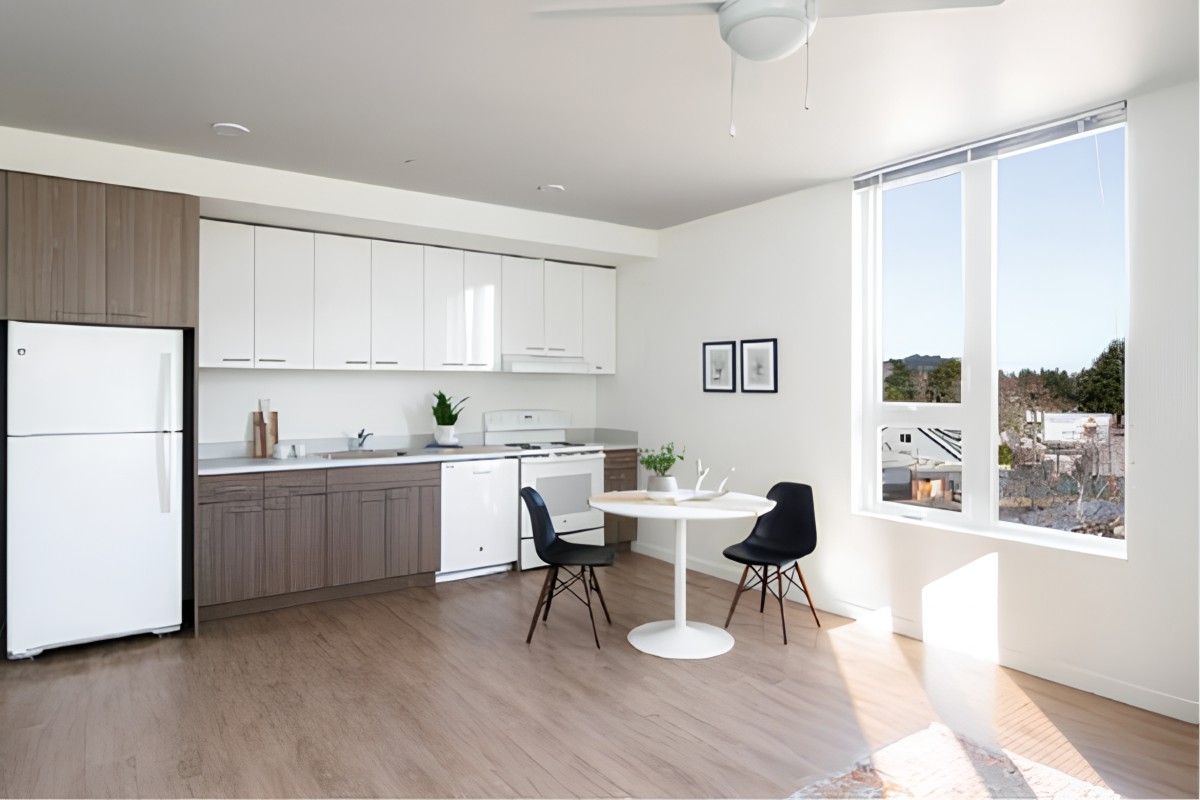 A modern and minimalist kitchen with a dining area, featuring white cabinets and natural lighting.