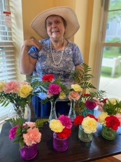 A smiling woman wearing a hat is surrounded by colorful flowers indoors.