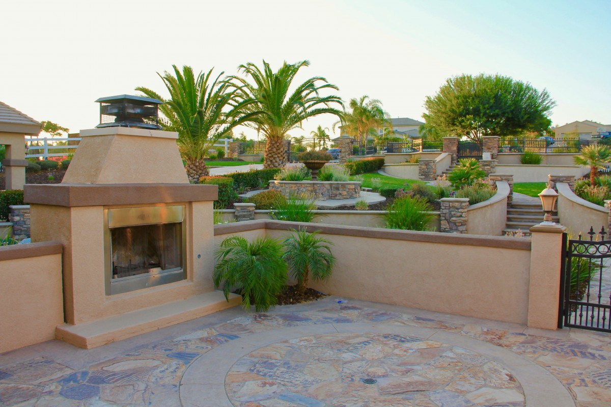 A luxurious outdoor patio featuring a stone fireplace, surrounded by lush landscaping and palm trees.