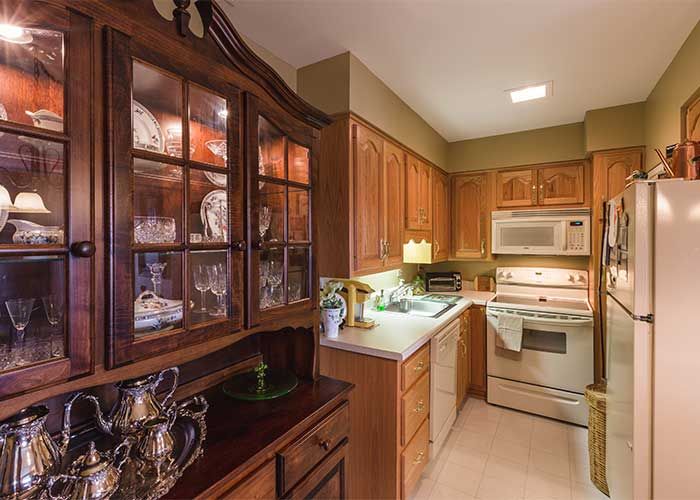A cozy kitchen with wooden cabinets, featuring a microwave and oven, and glassware displayed in a cabinet.