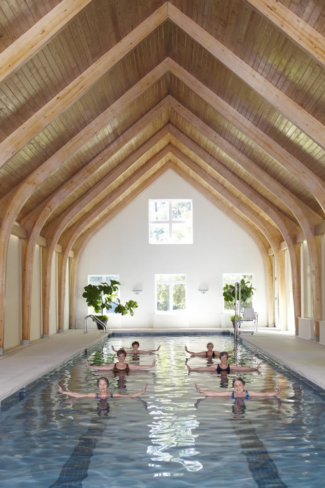 A group of people participating in a swimming exercise class inside a beautifully designed wooden structure with large windows and indoor plants.