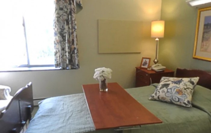 A hospital bedroom with a bedside table and lamp, featuring a vase of flowers and window curtains.